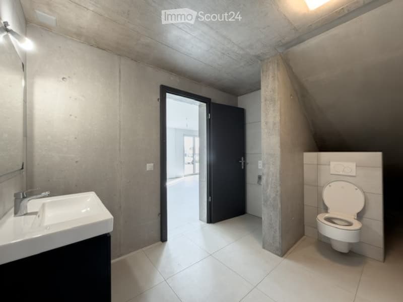 Modern bathroom with a white sink, chrome faucet, and toilet. The walls are concrete, and the floor is tiled.
