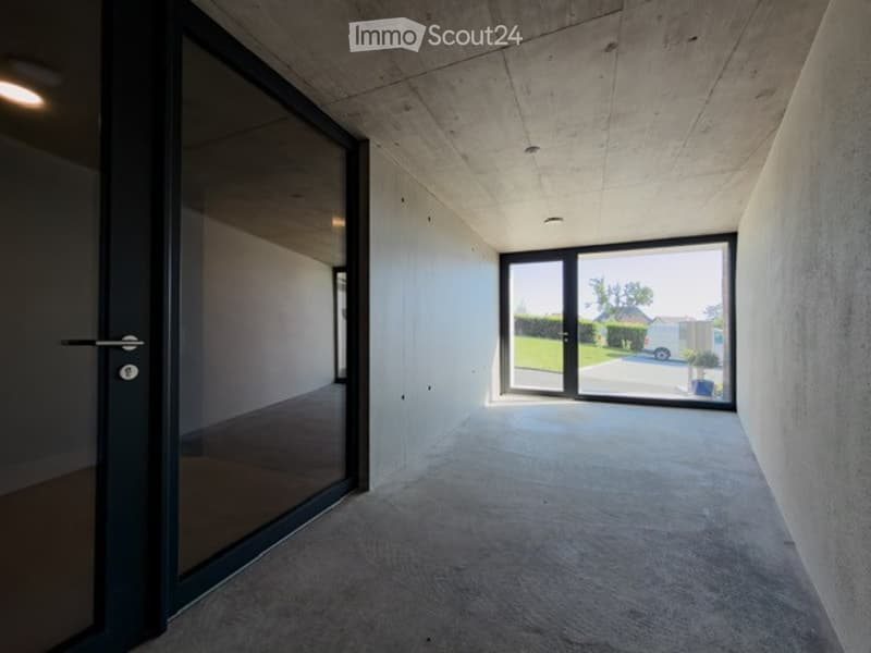Modern, minimalist hallway with concrete walls and floor, large glass doors leading outside, and recessed lighting.