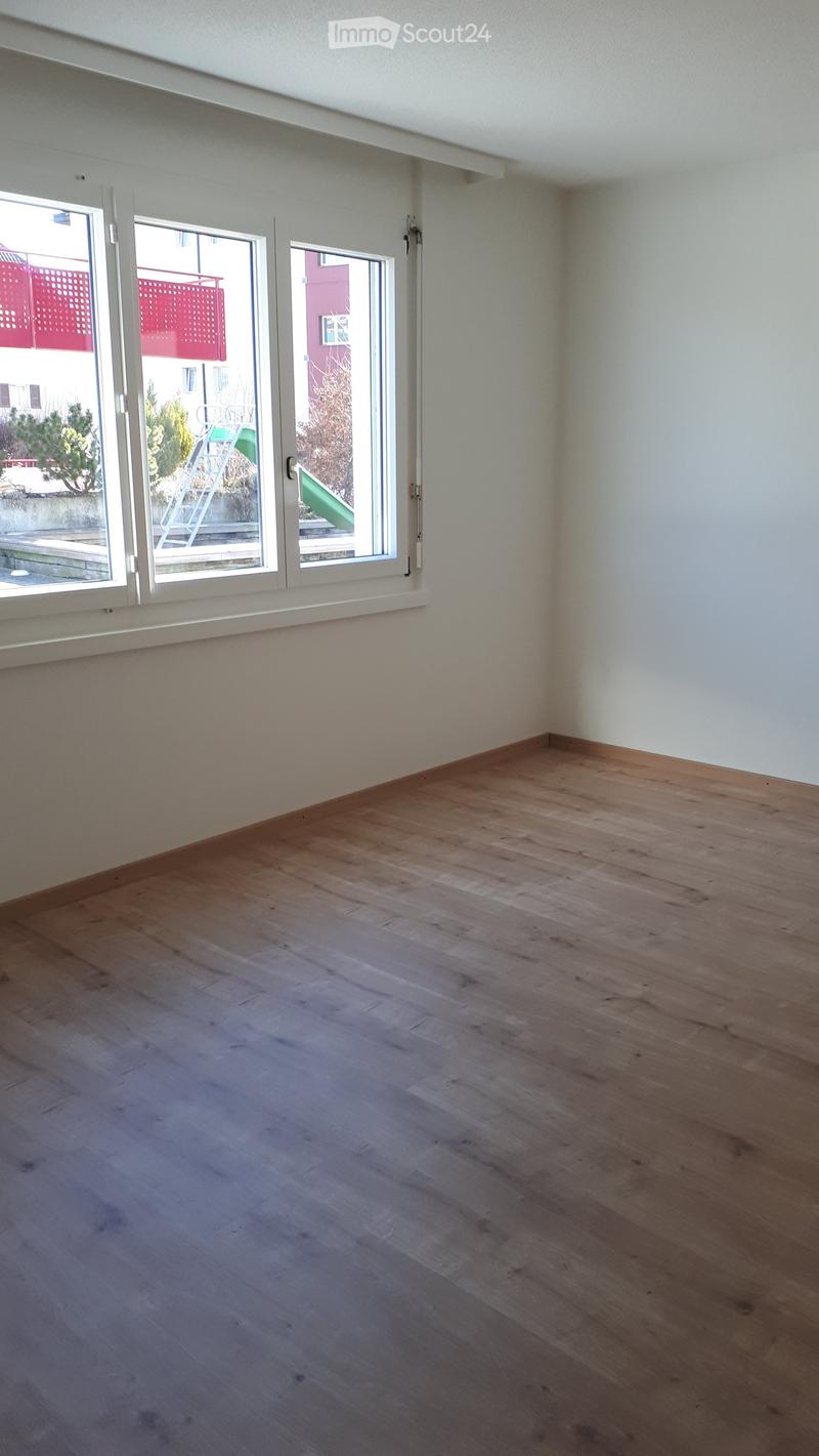 Empty room with wooden flooring and large windows, showing a view of the outside area.