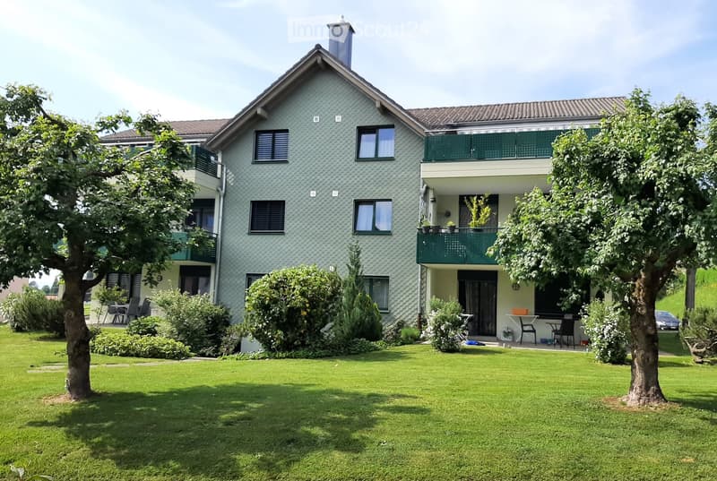 Two-story house with green siding, multiple windows, balconies, and a well-maintained lawn with trees and shrubs.