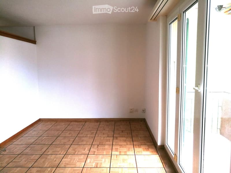Empty room with wooden parquet flooring, white walls, and large windows allowing natural light to enter.