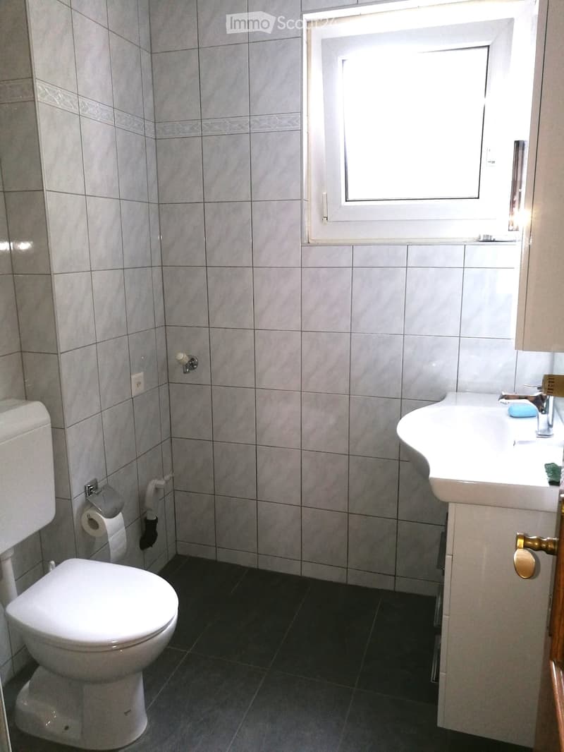 A small bathroom with a toilet, sink, and window. The walls are tiled in light gray with a decorative border near the top. The floor is dark gray tiles.
