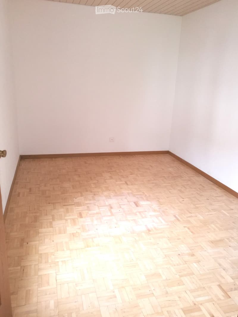 Empty room with light wooden herringbone flooring and white walls, featuring a visible electrical outlet and a door frame on the left side.