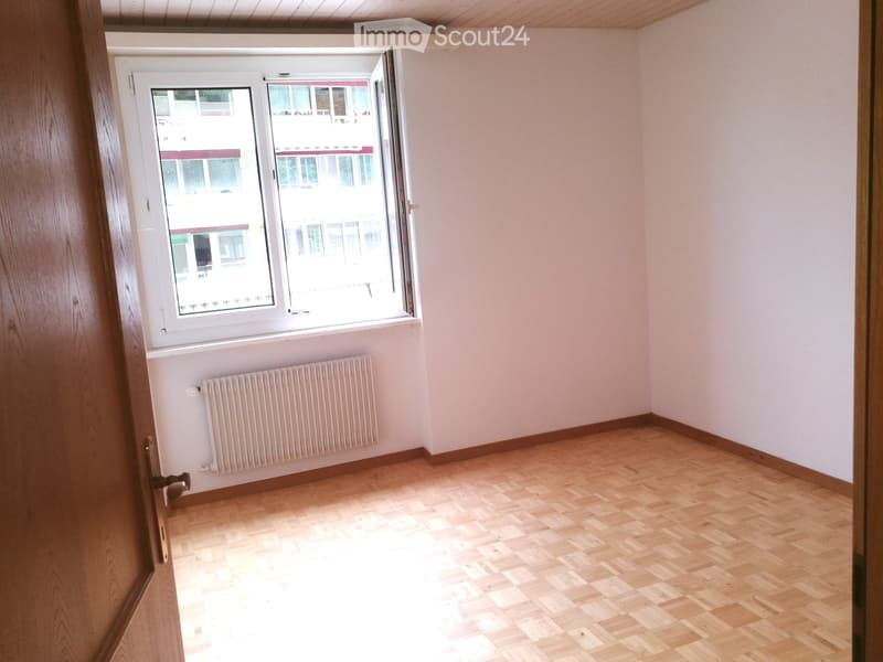 Empty room with wooden flooring, white walls, a radiator, and a large window with a view of a building outside.