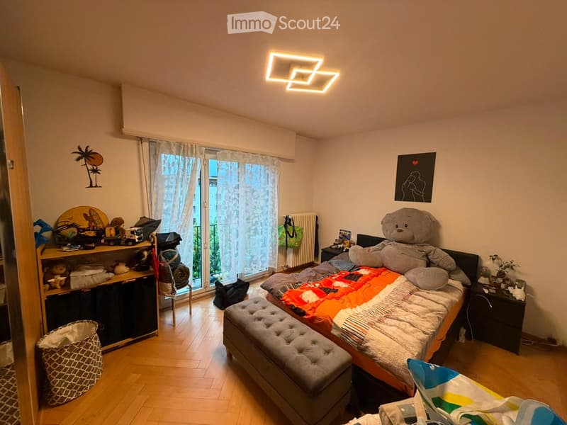 Cozy bedroom with wooden flooring, large window with curtains, bed with colorful bedding, nightstand, chair, and various decorative items including a large teddy bear and artwork.