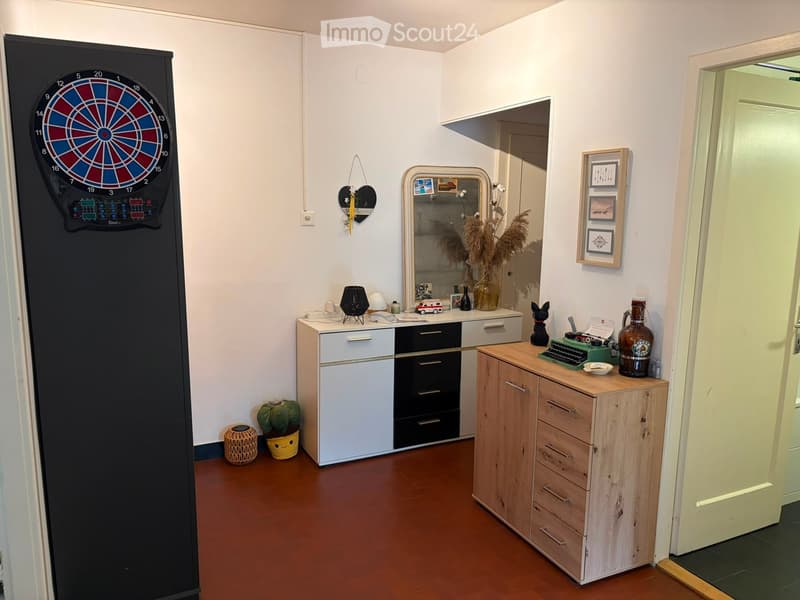 A small, modern room with a dartboard on one wall, a mirror, a desk with a lamp, a wooden cabinet, and various decorative items including framed pictures and a vase with dried flowers.