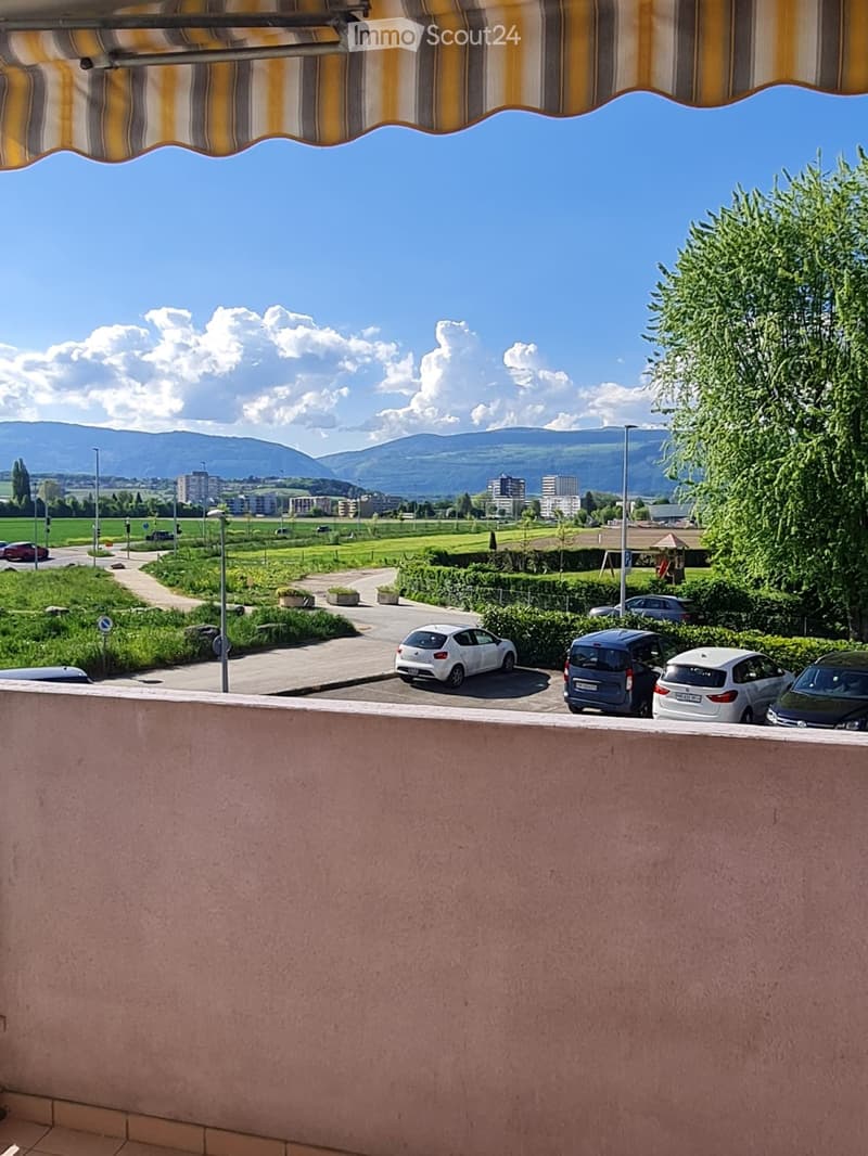 Outdoor balcony with a scenic view of the mountains, parking lot, cars parked, lush greenery, trees, and a clear blue sky