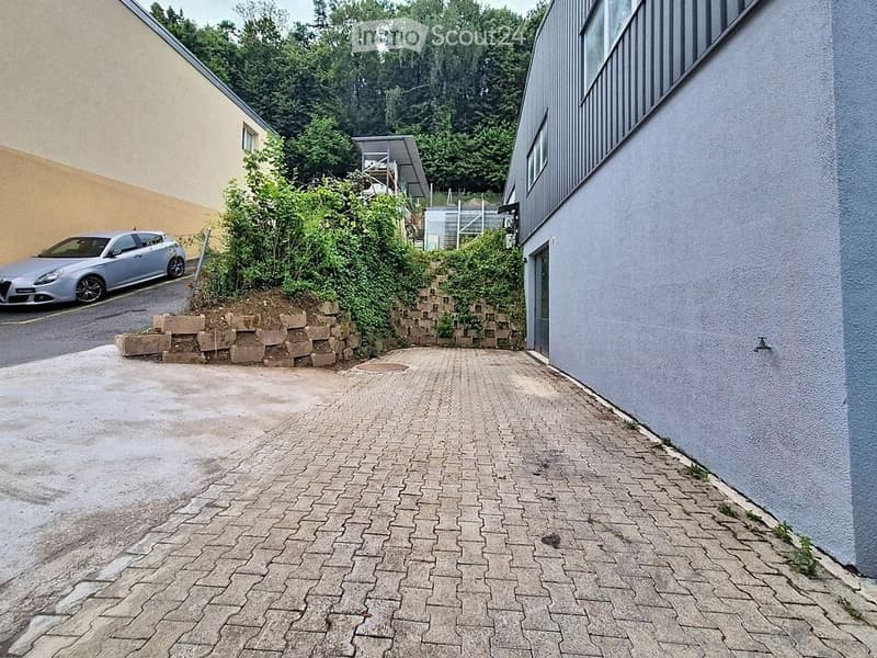 Brick-paved pathway leading to a modern building entrance, with a raised garden bed and a parked car visible in the background.