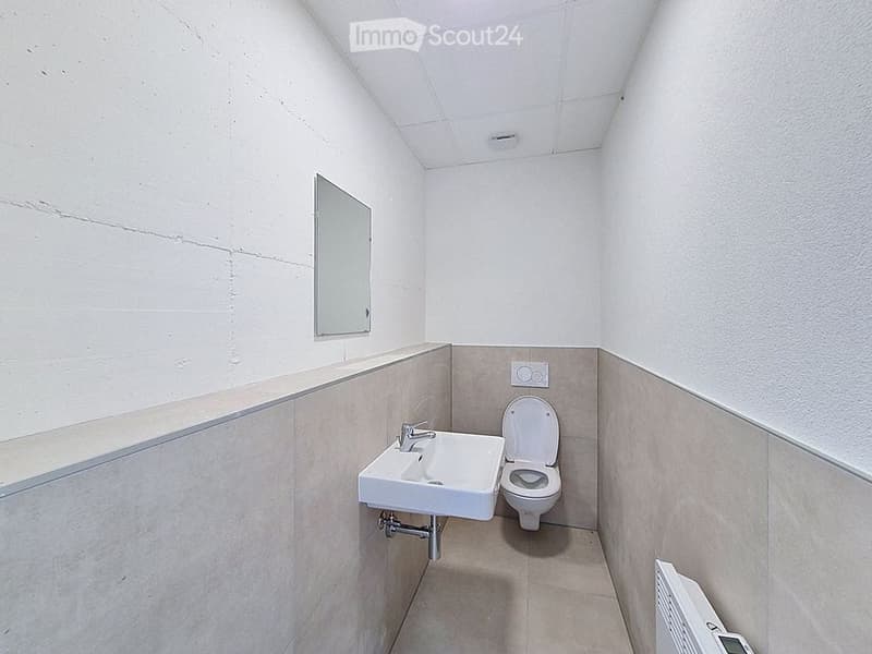 Small, modern bathroom with white walls, a white sink, a toilet, and a mirror. The floor and lower walls are tiled in a light beige color.