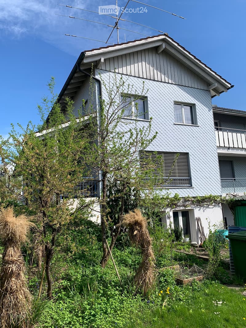 Two-story house with white siding, multiple windows, and a balcony on the upper level, surrounded by overgrown vegetation and a garden area.