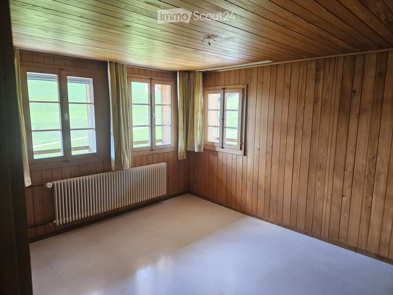 Empty room with wooden walls, wooden ceiling, tiled floor, and large windows with curtains. A radiator is visible on one wall.