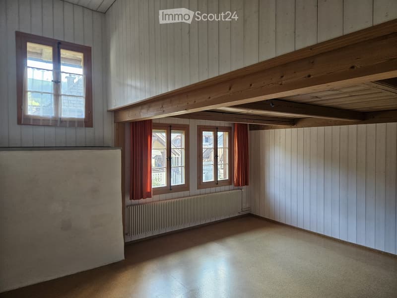 Empty room with wooden panel walls, wooden ceiling beams, and large windows with curtains. The floor is a light-colored concrete or tile surface.