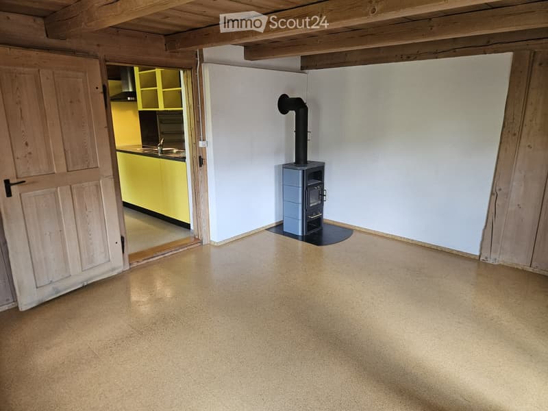 Empty room with exposed wooden beams, concrete floor, and a wood-burning stove with a chimney. A doorway leads to a brightly colored kitchen area.
