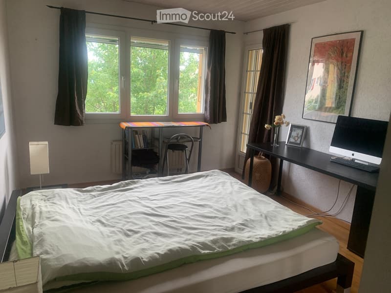 Bright bedroom with a double bed, desk, chair, lamp, and large windows with curtains. A vase with flowers and a framed picture are on the desk.