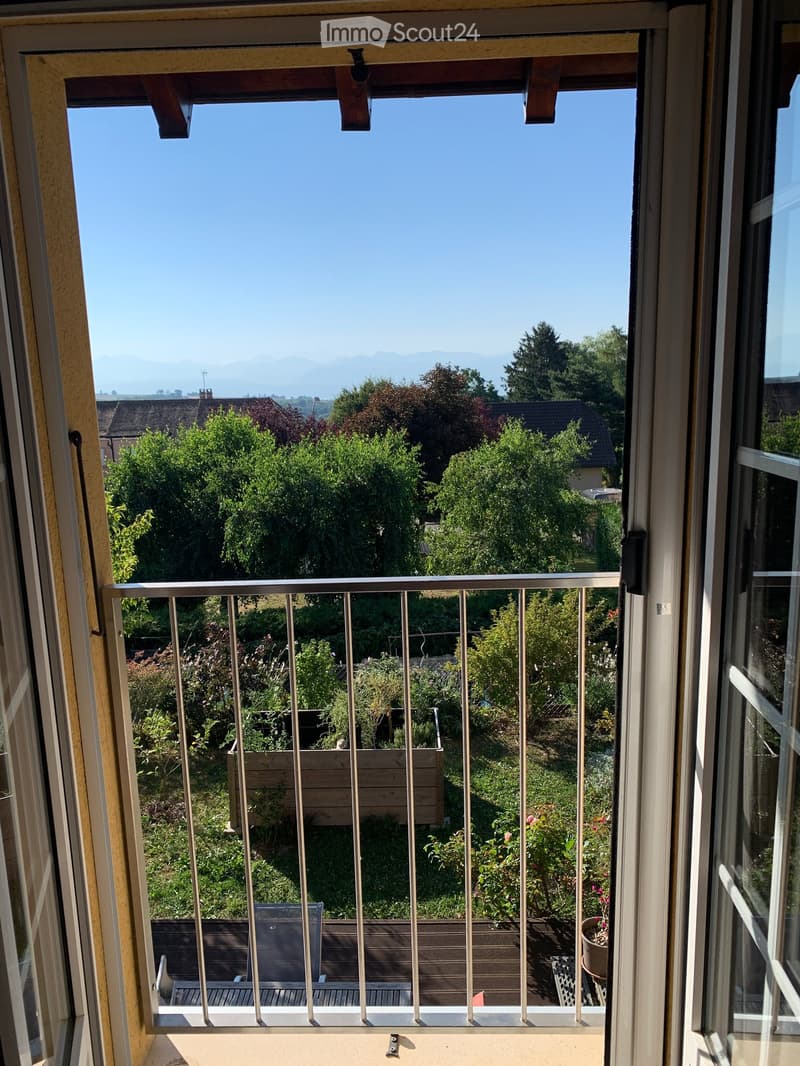 Balcony with metal railing, overlooking a garden area with plants, trees, and distant mountains under a clear blue sky.