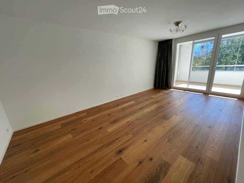Empty room with wooden flooring, white walls, large sliding glass door leading to a balcony, and a chandelier on the ceiling.