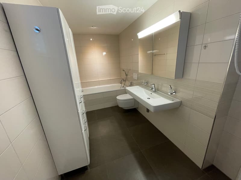 Modern bathroom with a white sink, toilet, bathtub, and a large mirror with built-in lighting. The walls are tiled in light beige, and the floor is dark grey.