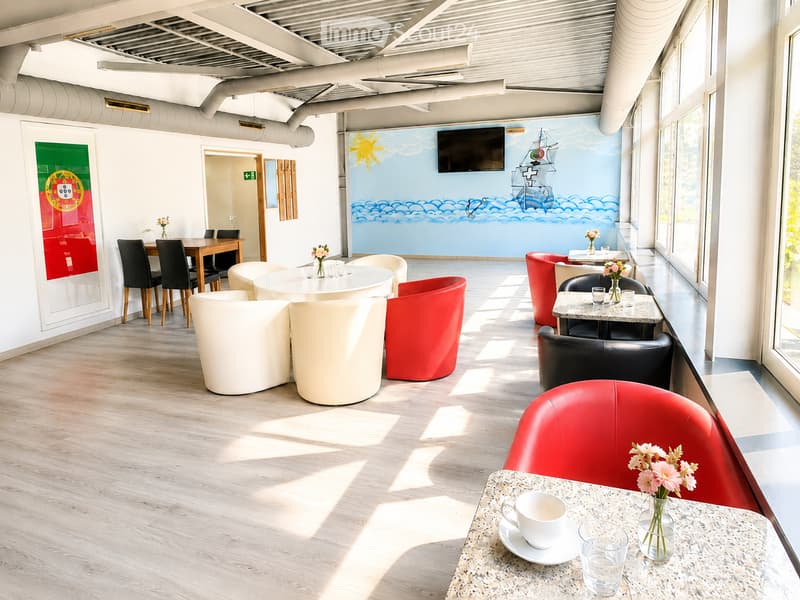Brightly lit room with wooden flooring, white and red chairs, tables, and a mural of a ship on the wall. There is a television mounted on the wall and a vase with flowers on a table.