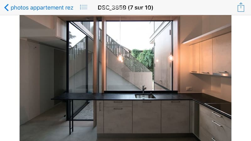 Modern kitchen with sleek, light-colored cabinets, a black countertop, a sink, and a small table near large windows offering a view of the outdoor area.