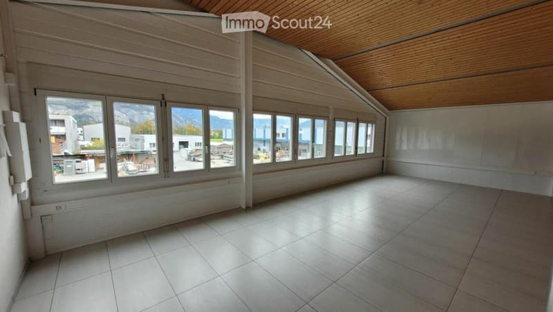 Empty room with large windows offering a view of the surrounding area, including buildings and mountains. The room features a tiled floor and wooden ceiling.