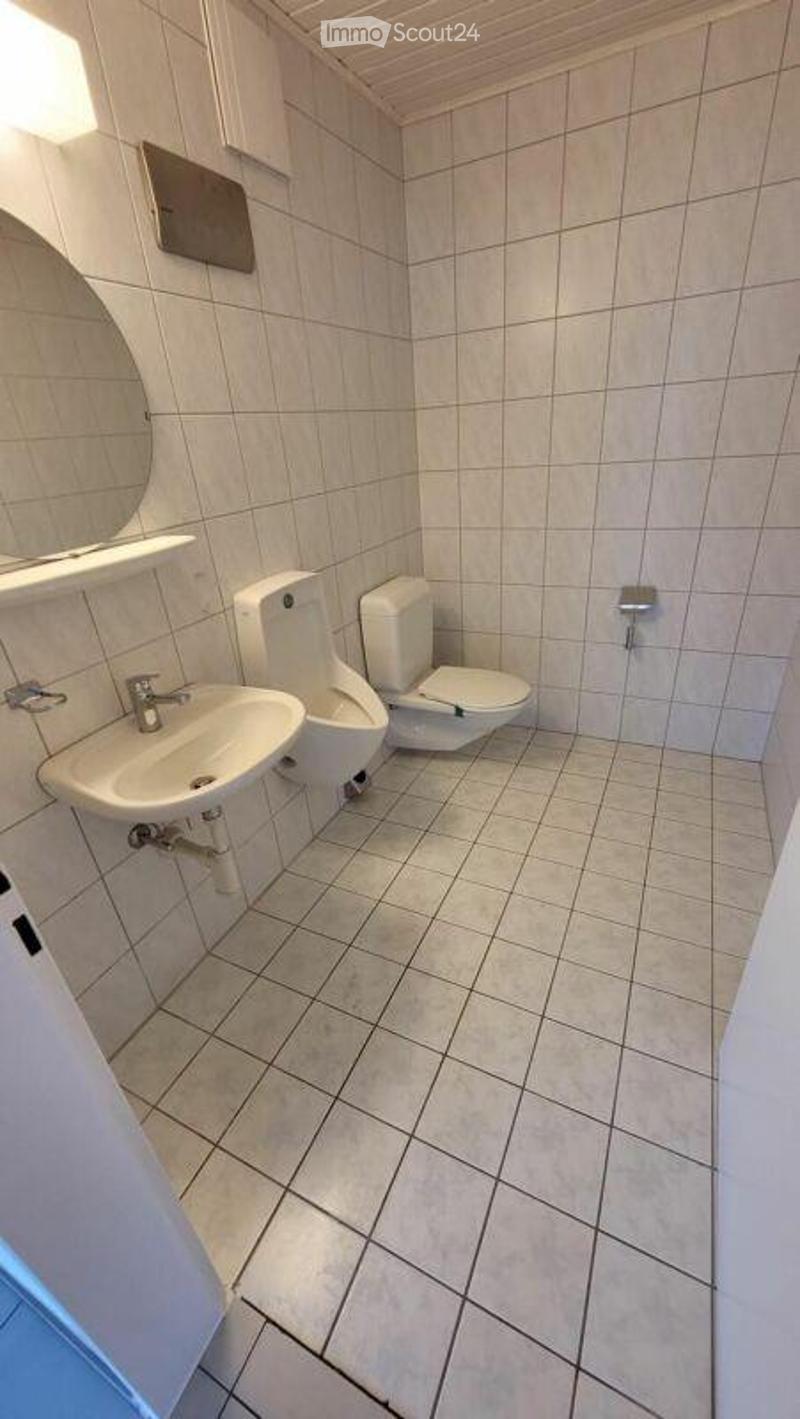 A small, clean bathroom with white tiled walls and floor, featuring a sink, toilet, urinal, and a mirror above the sink.