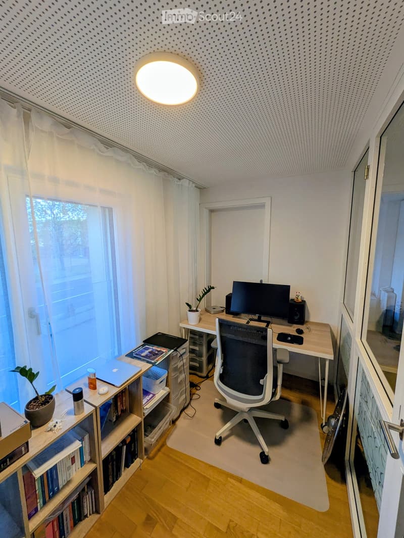 A modern, well-lit study or home office with a wooden desk, ergonomic chair, laptop, monitor, bookshelf, and decorative plants.