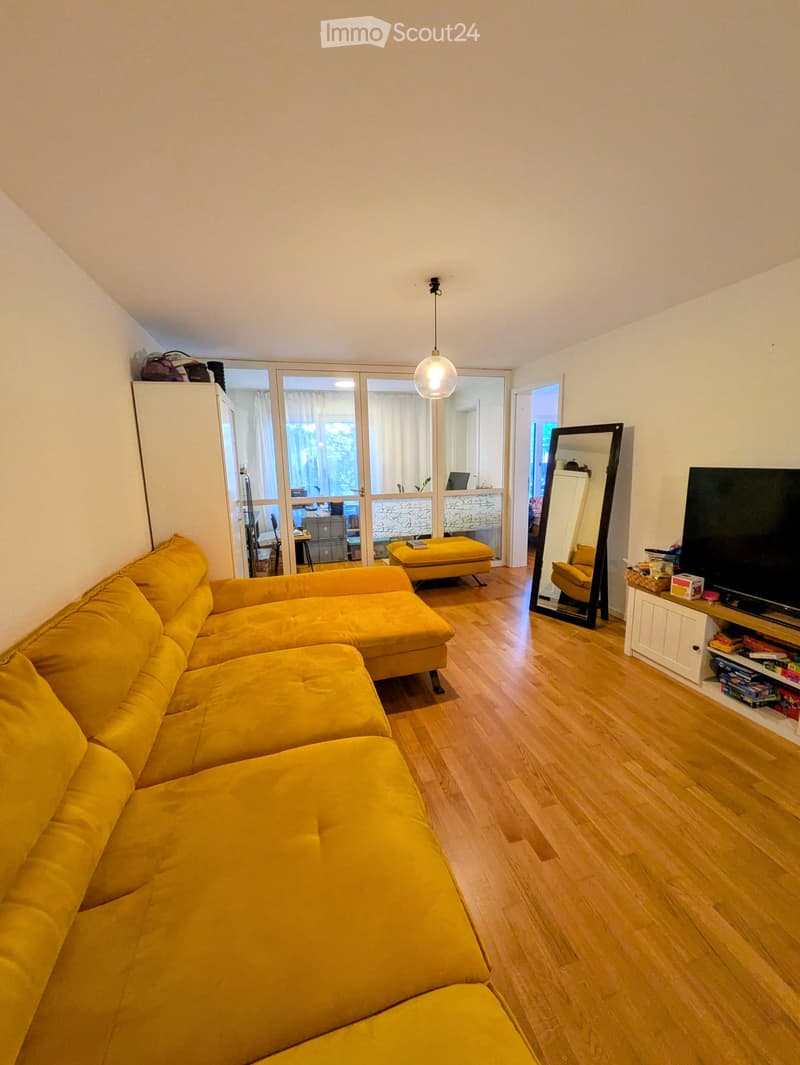 Living room with a yellow sectional sofa, ottoman, large mirror, television, and a small table with a chair visible through the open door.