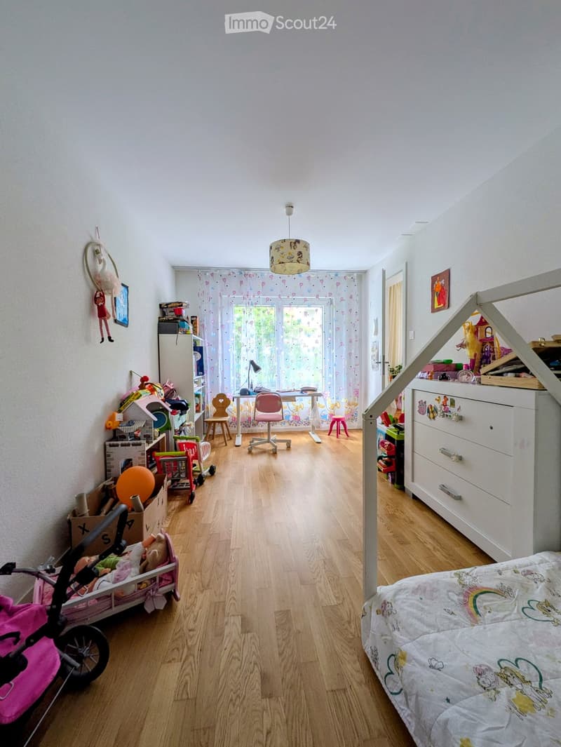 A well-lit children's bedroom with wooden flooring, a bed with a colorful blanket, a desk with a chair, a small table with a lamp, a refrigerator, and various toys scattered around, including a toy car, a toy house, and a toy stroller.