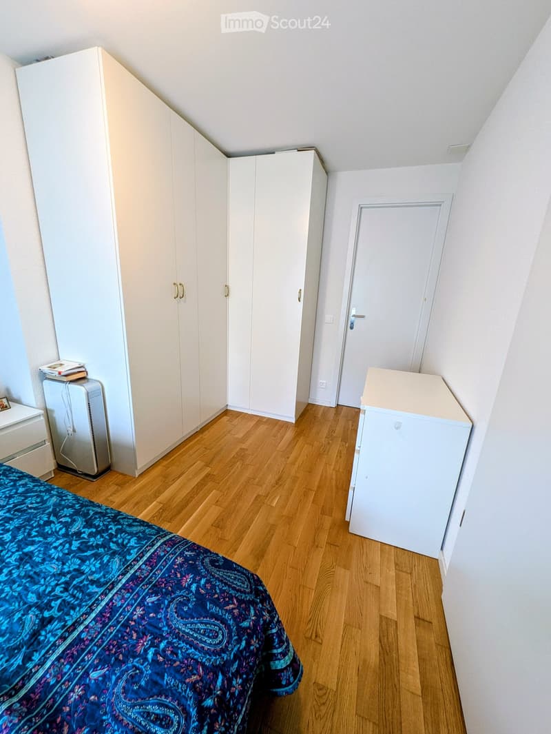 A well-lit bedroom with wooden flooring, a large bed with a blue patterned bedspread, a white wardrobe, a small refrigerator, and a nightstand with books.