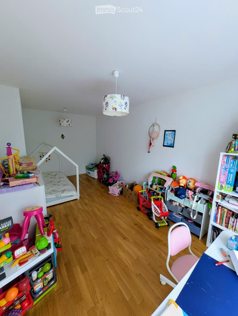 A well-lit children's room with wooden flooring, a bed, a chair, a table, shelves filled with toys, and a colorful play area.