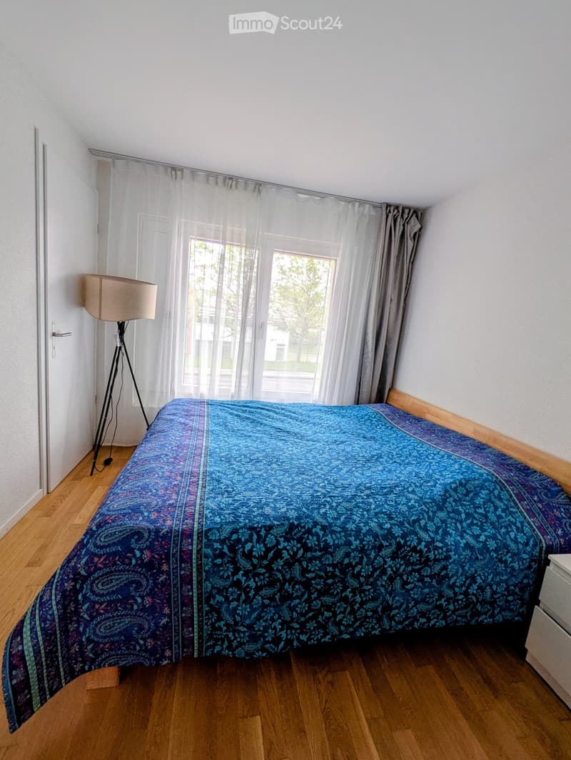 Bedroom with wooden floor, large window with sheer curtains, bed with blue patterned duvet, floor lamp, and nightstand.