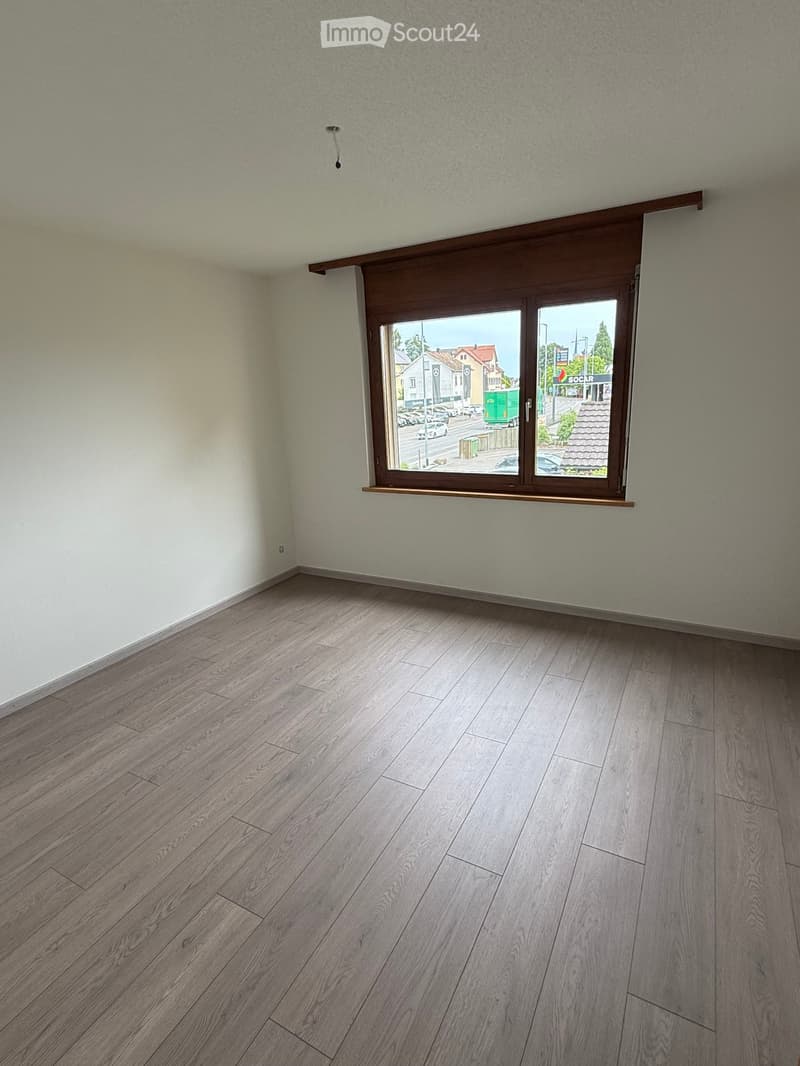 Empty room with light-colored walls and wooden flooring, featuring a large window with a brown frame that offers a view of the street and surrounding buildings.