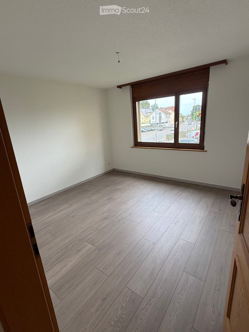 Empty room with light grey wooden flooring and white walls, featuring a large window with wooden frame and blinds, overlooking a street with parked cars and buildings.
