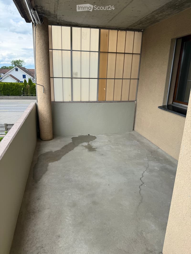 Empty balcony with concrete floor, beige walls, and a metal railing with a wooden door leading outside.