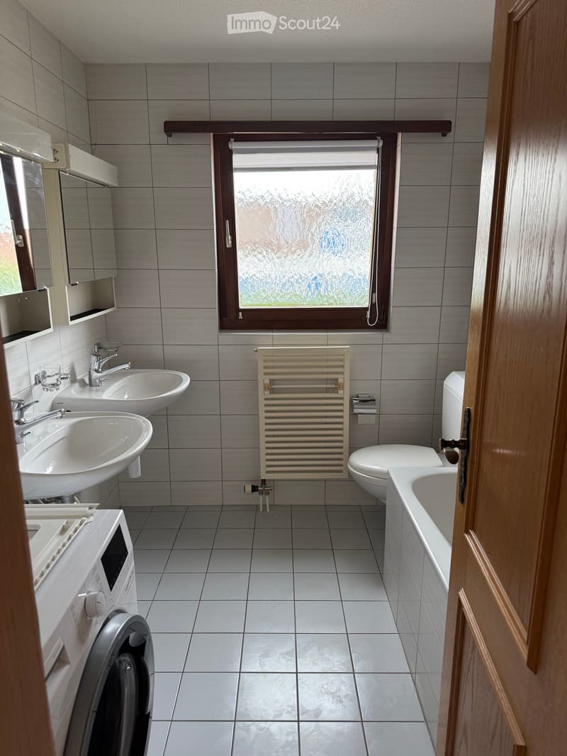 Modern bathroom with two sinks, a toilet, a bathtub, a radiator, a mirror, and a washing machine. The room features grey tiles and a frosted window.