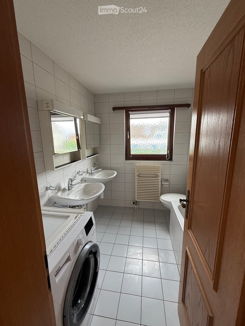 Modern bathroom with two sinks, a mirror, a radiator, a toilet, and a washing machine. The room features tiled walls and floor, with a window providing natural light.