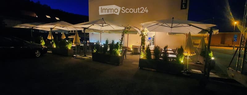 Outdoor seating area with tables, chairs, and large umbrellas, surrounded by potted plants and illuminated lighting.
