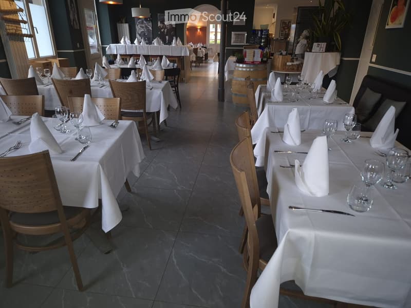 Restaurant interior with multiple tables set for dining, each with white tablecloths, wine glasses, napkins, and cutlery. Wooden chairs surround the tables, and the space features large windows and a barrel in the background.