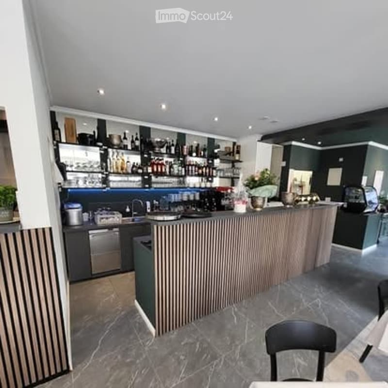 Modern kitchen with a bar counter, shelves filled with bottles and glasses, a sink with a faucet, and a dishwasher. A black chair is placed near the counter.