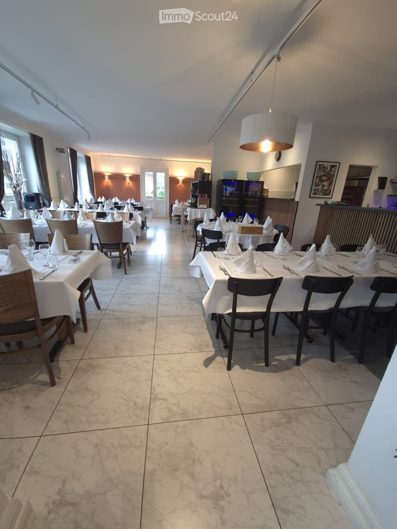 Restaurant dining area with multiple tables set for guests, featuring white tablecloths, napkins, cutlery, and glasses. Wooden and black chairs are arranged around the tables. A bar area with a refrigerator and picture frames is visible in the background.