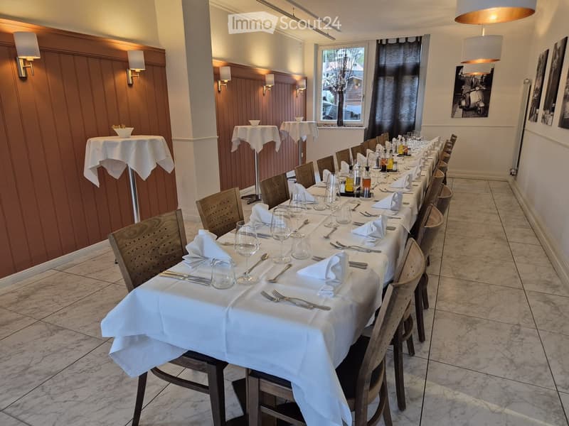 Elegant dining area with long table set for multiple guests, featuring wine glasses, cutlery, and napkins, surrounded by wooden chairs and additional smaller tables.