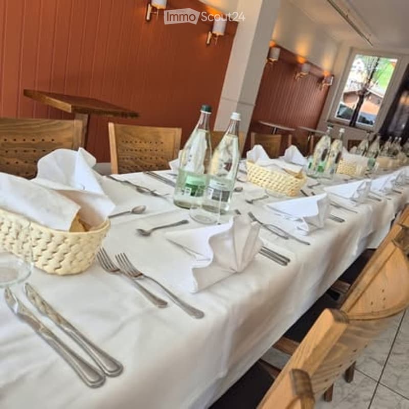 A long dining table set for a formal meal, with multiple place settings, bread baskets, and water bottles. Wooden chairs surround the table, and the room has warm wooden paneling and large windows.