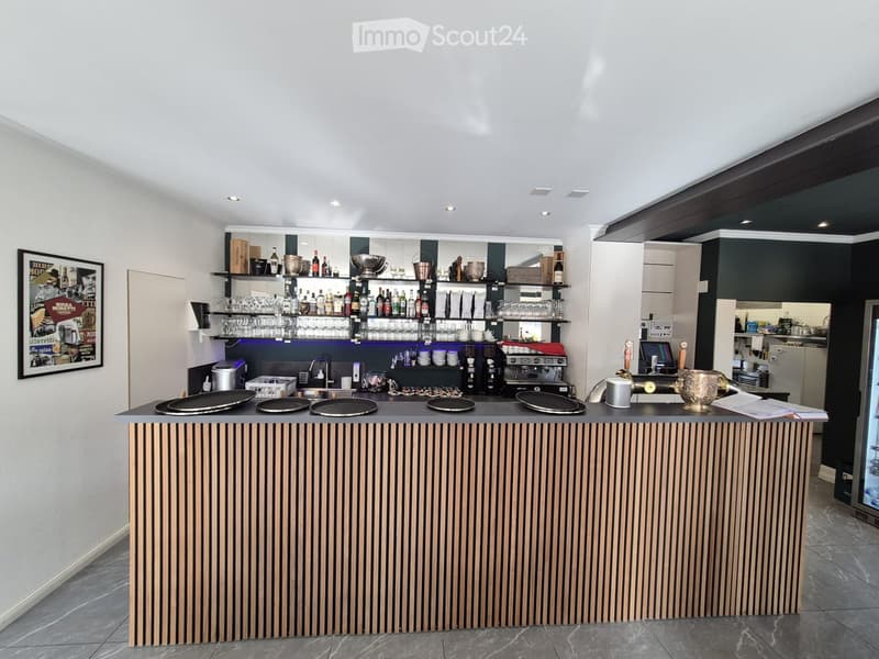 Modern bar area with a wooden counter, shelves filled with bottles and glasses, a coffee machine, and a refrigerator. The space is well-lit with recessed lighting and features a sleek, contemporary design.