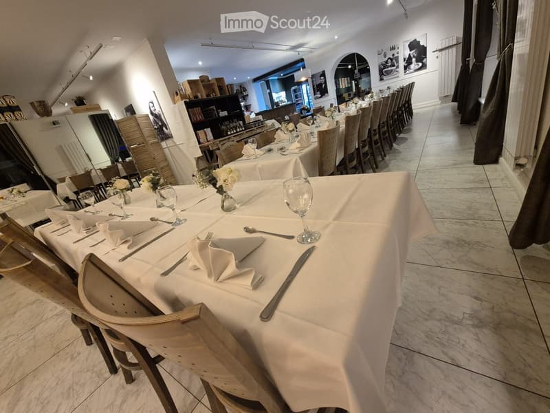 Restaurant dining area with long tables, chairs, and place settings, featuring white tablecloths, silverware, and decorative vases.