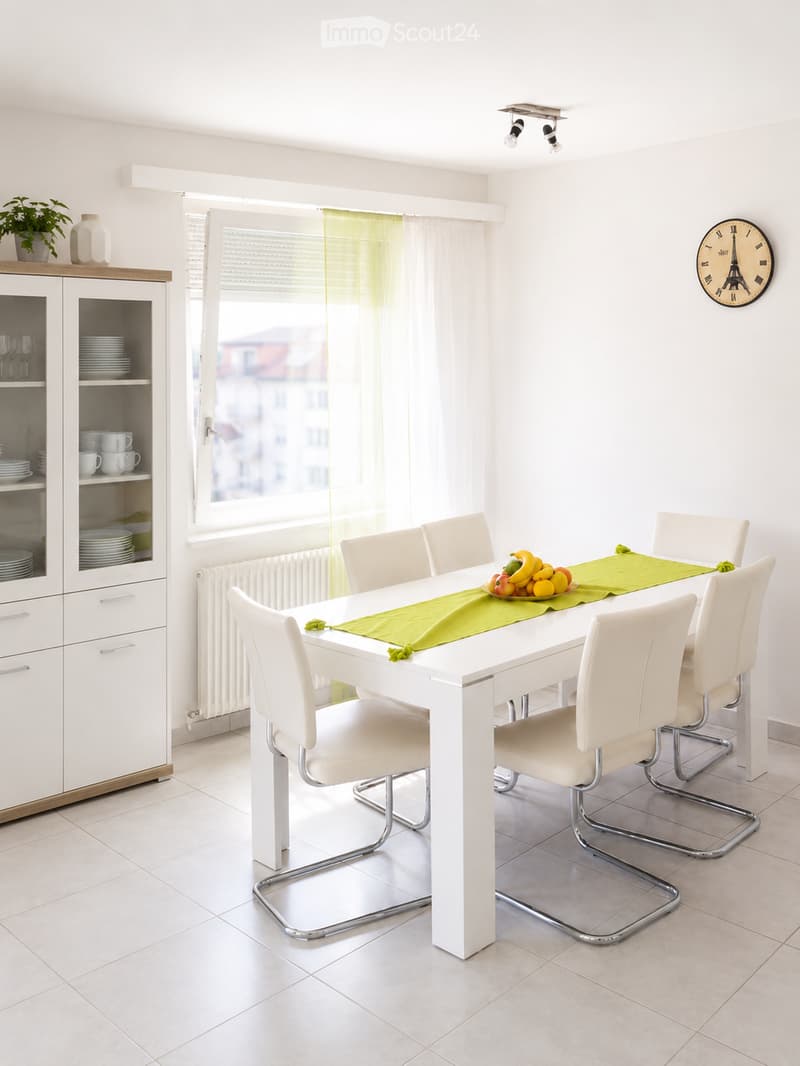 Bright dining room with white dining table, six white chairs, green table runner, fruit bowl, white cabinet with glass doors, and a clock on the wall.