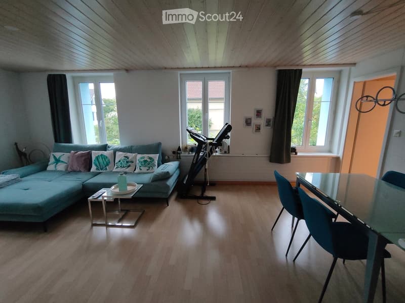 Bright living room with a teal sectional sofa, coffee table, exercise bike, and dining area with a glass table and chairs.