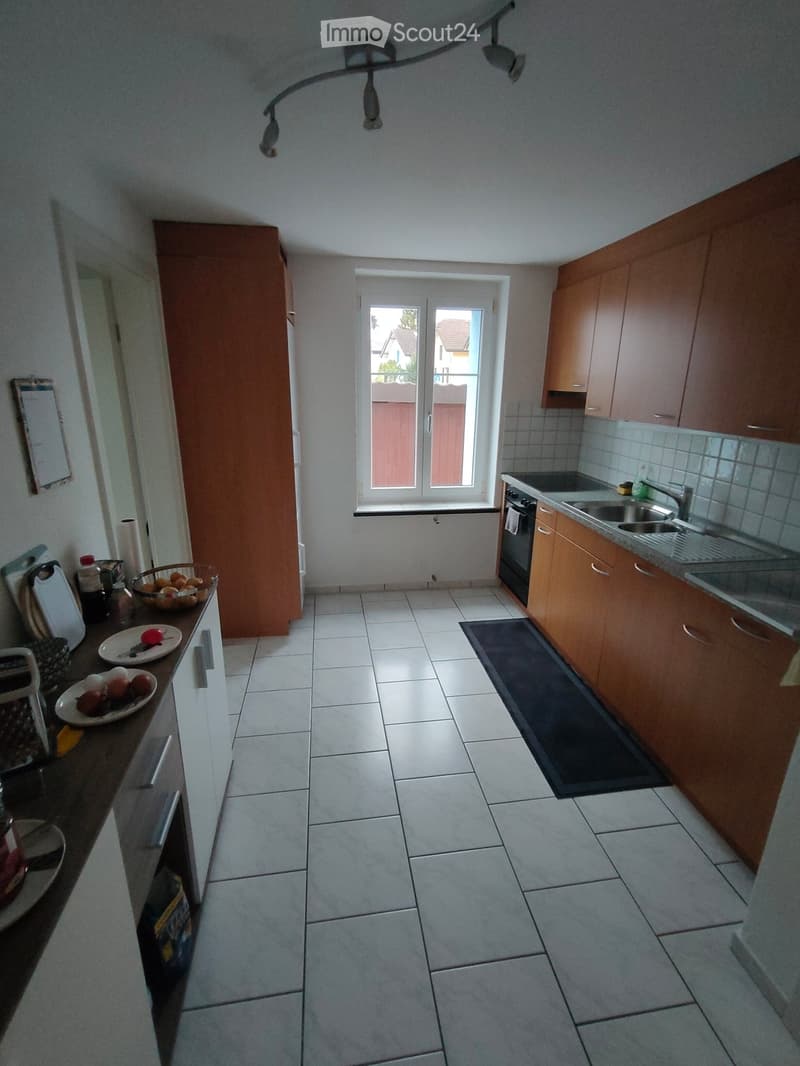 Modern kitchen with wooden cabinets, tiled floor, and stainless steel appliances including an oven, sink, and faucet. Countertop has plates with fruit and a bowl of eggs.