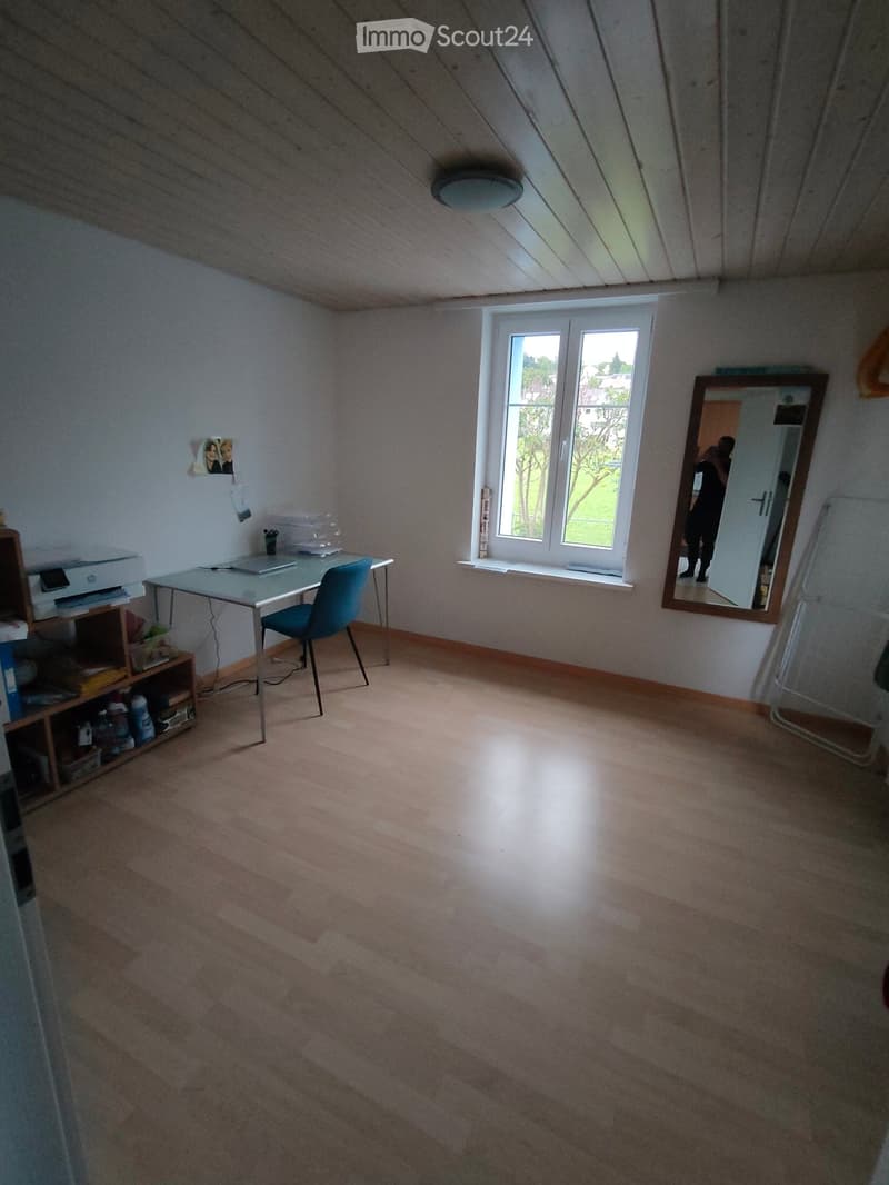 Bright room with wooden floor, large window, small table with chair, shelf with items, and mirror on wall.