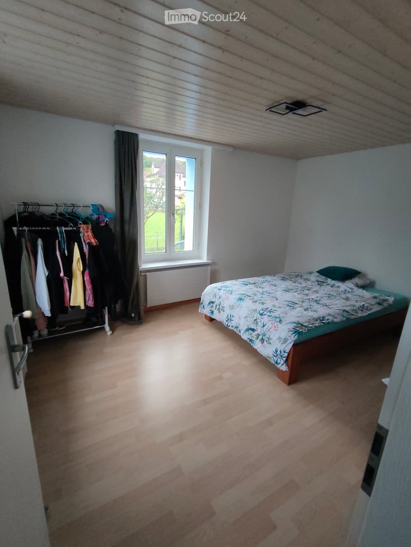 Bedroom with wooden floor, bed with floral bedding, wardrobe with hanging clothes, large window with view.