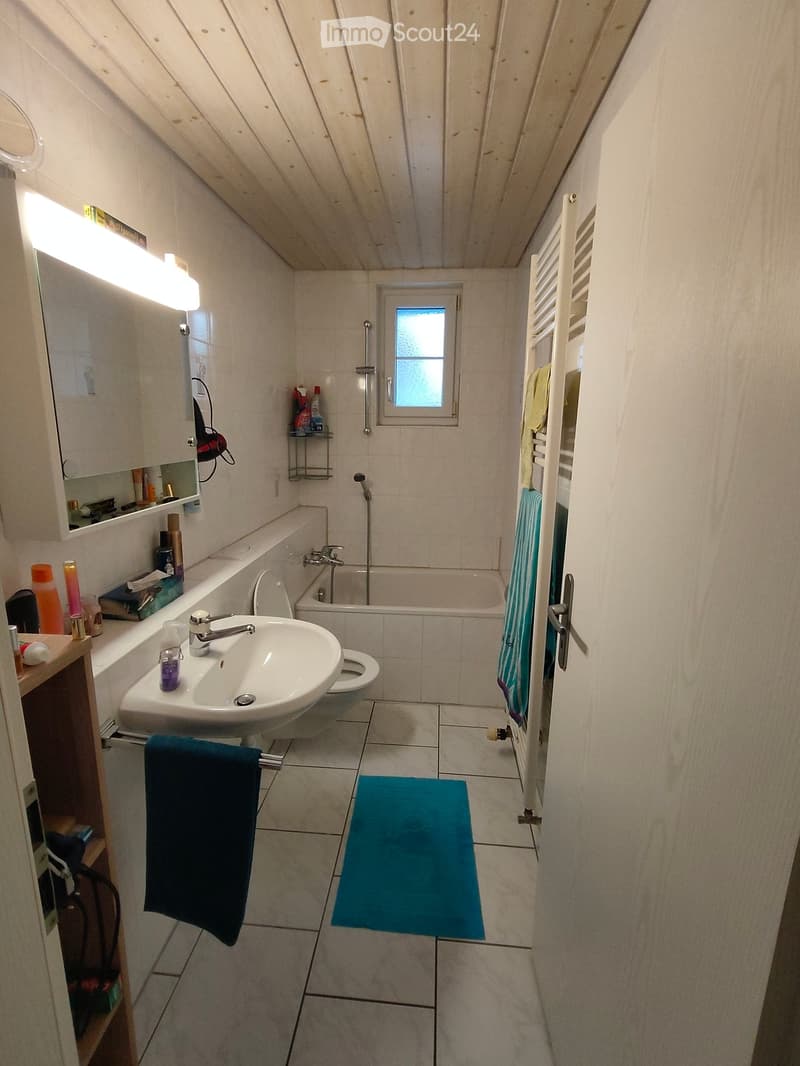 Bathroom with a sink, toilet, bathtub, mirror, towel, bath mat, and a small window. The room features tiled walls and floor, and a towel rack with towels.